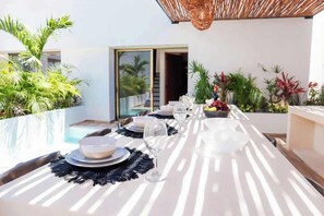Un espace de restauration en plein air avec une table dressée pour deux personnes, entouré d’une végétation luxuriante et offrant une vue sur une piscine.