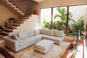 Un salon moderne avec un canapé d’angle, un petit pouf, un escalier doté d’étagères intégrées et une grande fenêtre donnant sur un jardin.