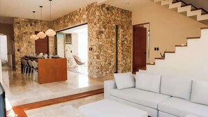 A modern living room with a stone accent wall, a white sofa, and a dining area with a wooden table and chairs. - Huge 10BR Luxury Villa Complex w/ 6 Private Pool (Tulum)
