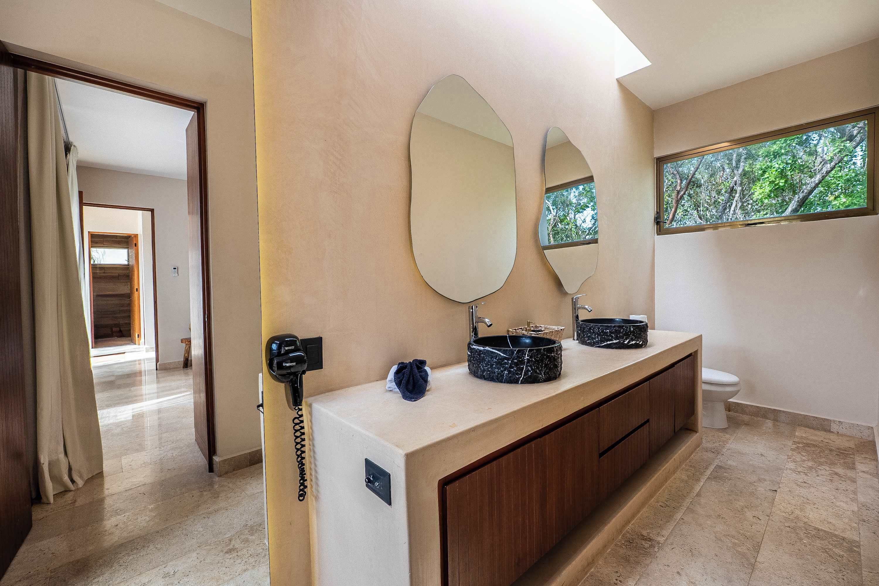 A modern bathroom with a vanity, two mirrors, a sink with a basin, a telephone, and a window with a view of trees.