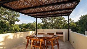 Un patio techado al aire libre con una mesa redonda de madera y sillas, una parrilla y vistas a árboles.