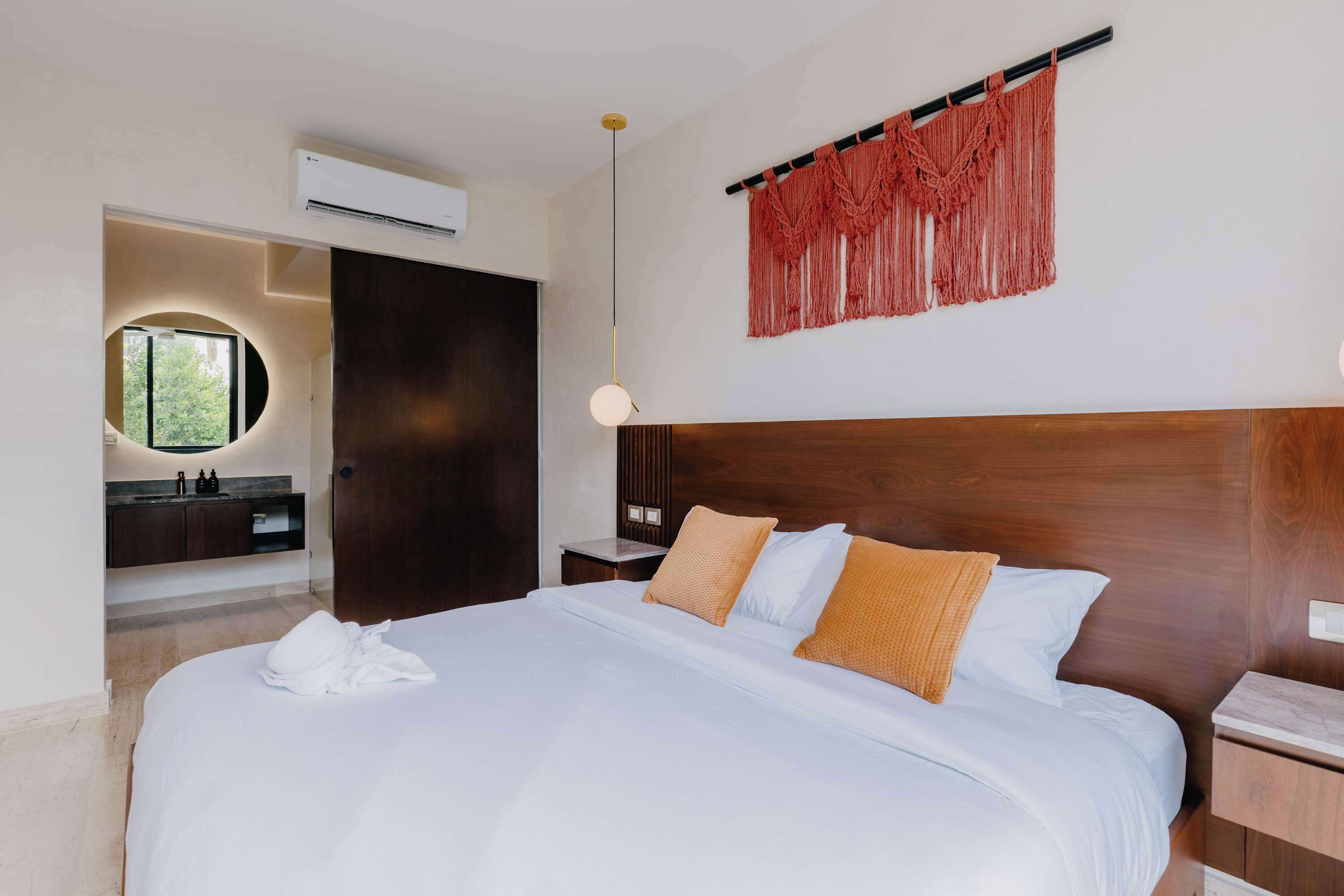 Habitación de hotel con una cama grande, cabecera de madera, espejo redondo, tocador con lavamanos y un colgador de macramé en la pared.
