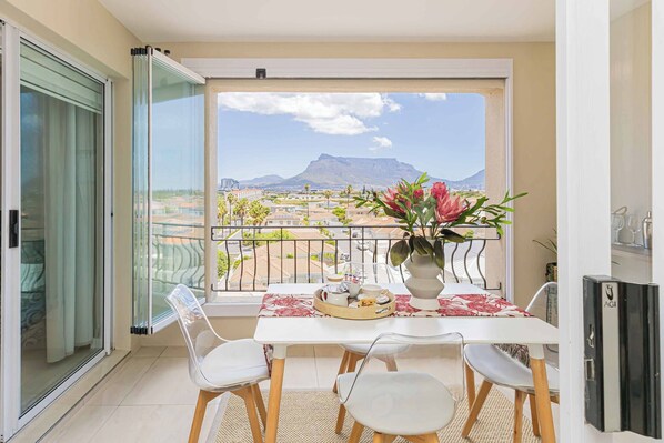 A balcony with a table set for two, a vase with flowers, and a view of a mountain and buildings. - Bella View of Magestic Table Mountain (Cape Town)