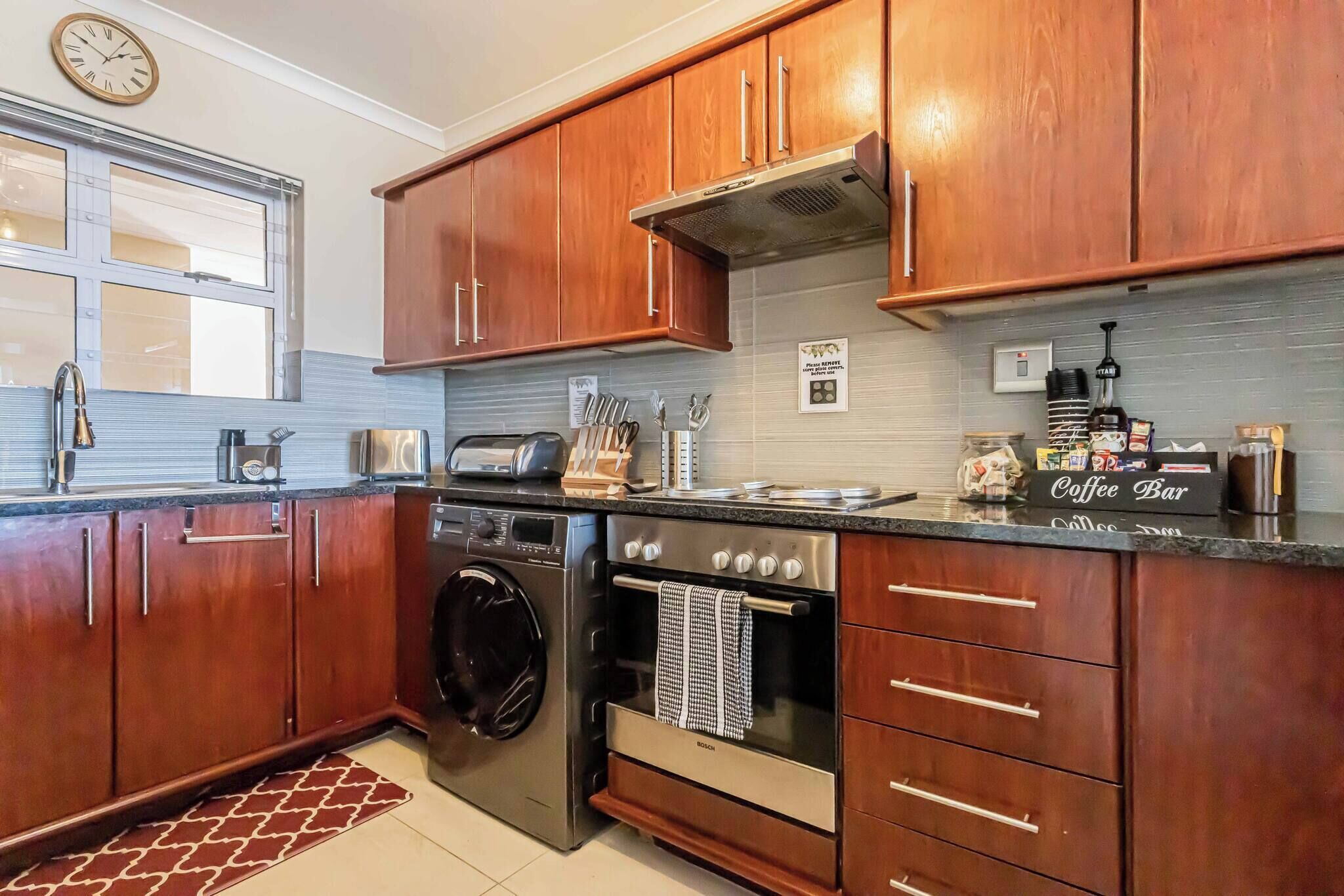 A kitchen with wooden cabinets, a Bosch washing machine, a coffee bar, and a clock on the wall.