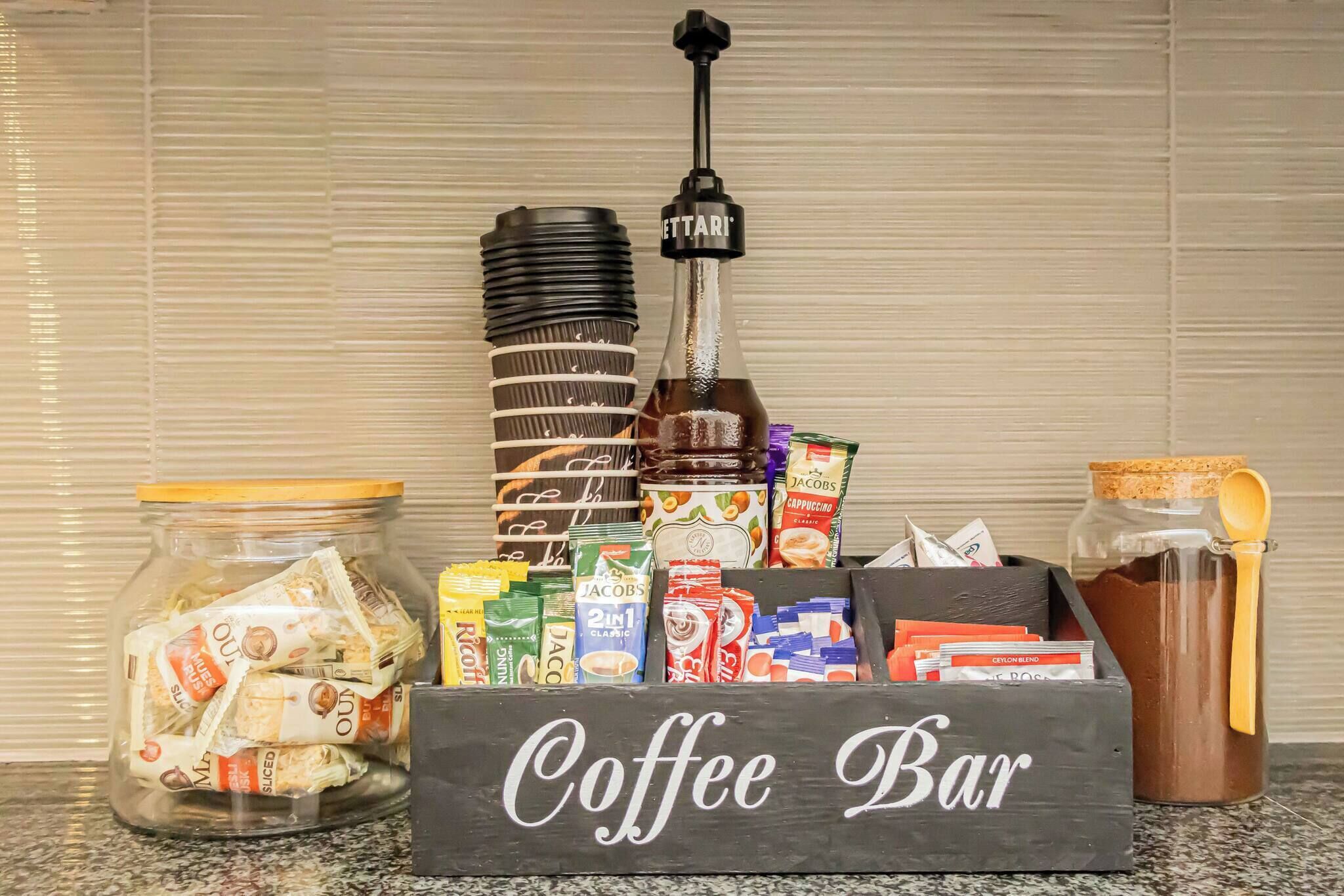 Uma barra de café com moedor, diferentes cápsulas de café, um frasco de açúcar e uma caneca.