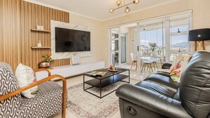 A modern living room with a flat-screen TV, leather sofa, patterned armchair, coffee table, and a view of the city through large windows. - Bella View of Magestic Table Mountain (Cape Town)