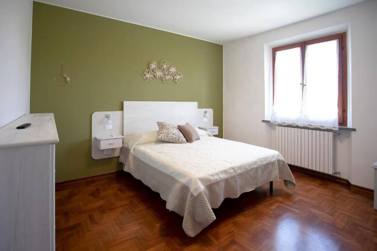 A bedroom with a bed, a nightstand, a wall-mounted lamp, a radiator, and a window with curtains.