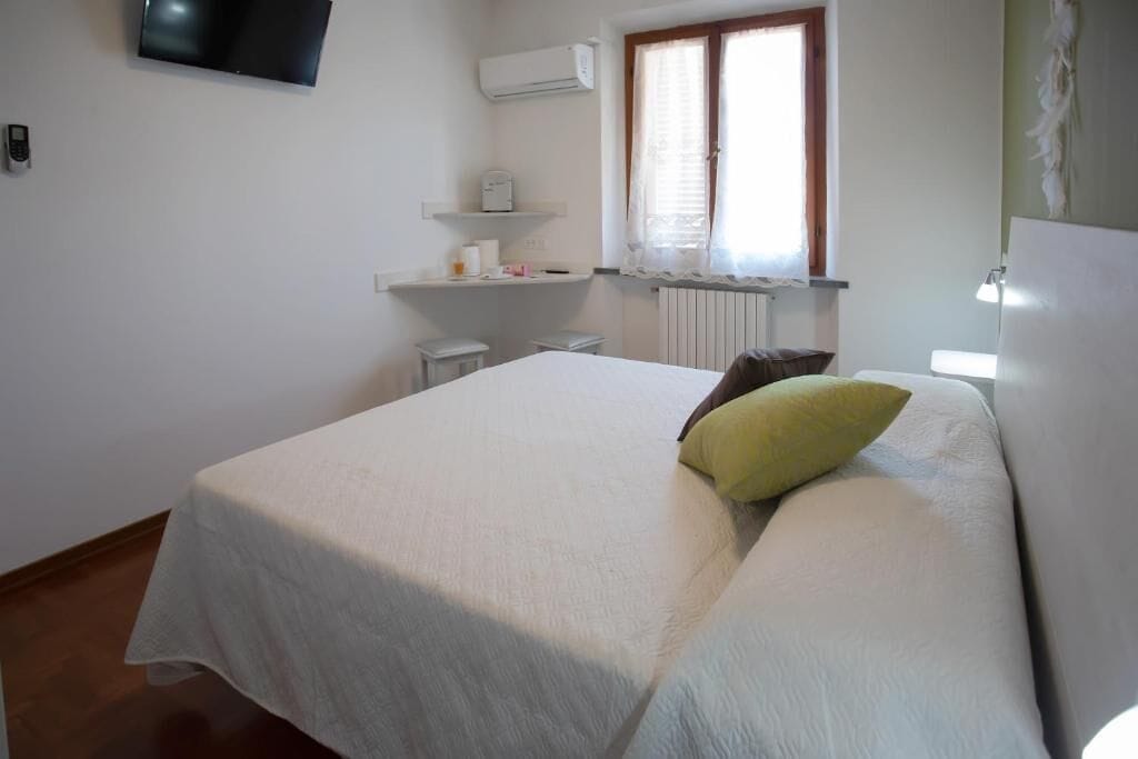 Un dormitorio con cama, televisor montado en la pared, ventana con persianas, radiador y una pequeña mesa con silla.