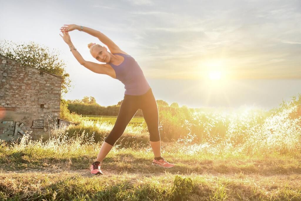 A person stretching outdoors with a stone wall and greenery in the background.