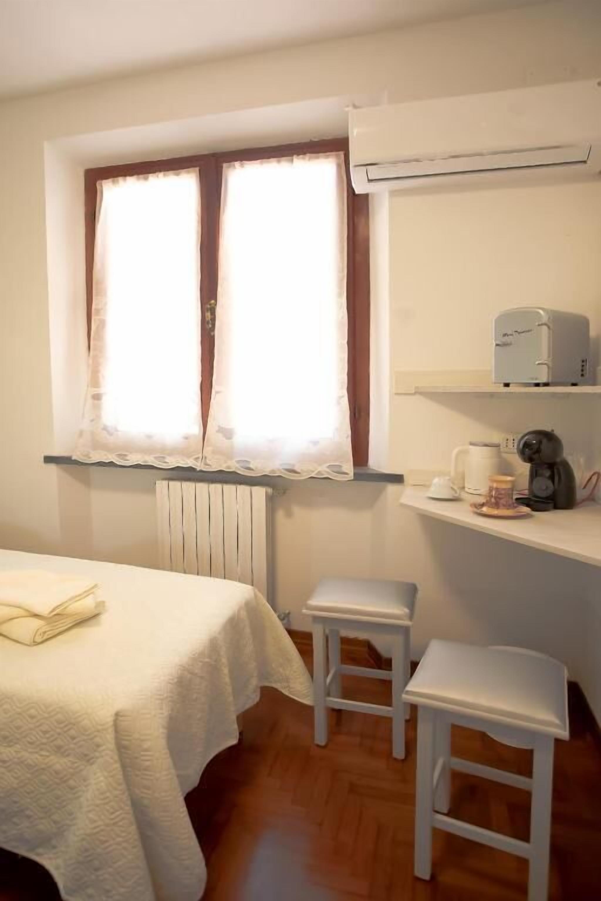 A small room with a bed, two white stools, a radiator, a window with sheer curtains, and a shelf with a coffee maker and other items.