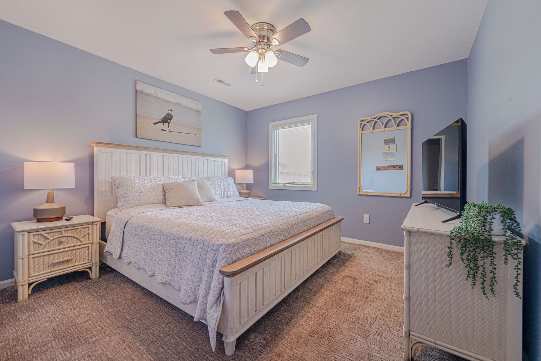 A bedroom with a bed, nightstands, a ceiling fan, a window, a mirror, and a plant.