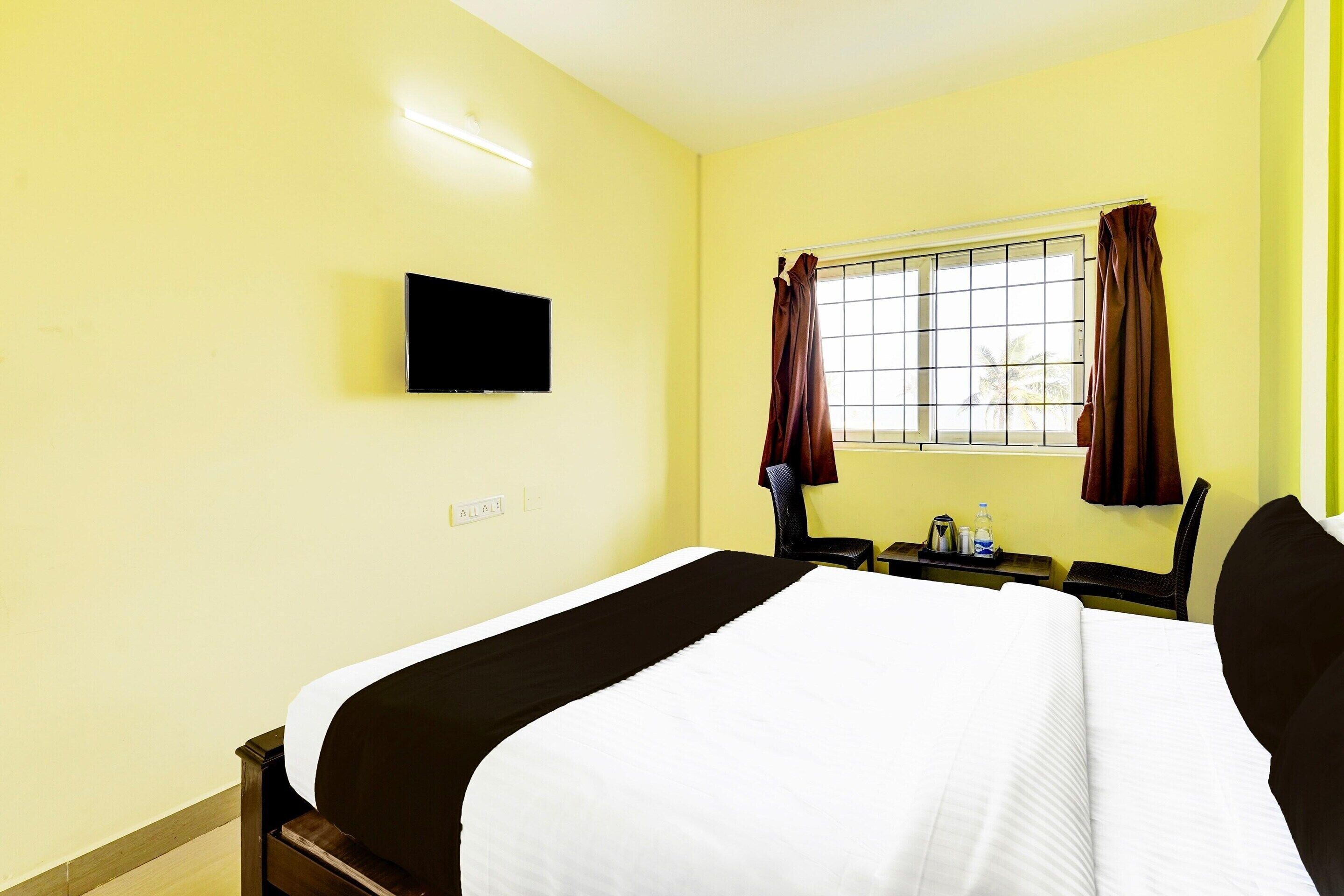 Habitación de hotel con una cama individual, un televisor de pantalla plana montado en la pared, una ventana con cortinas y una pequeña mesa con una botella de agua y una taza.