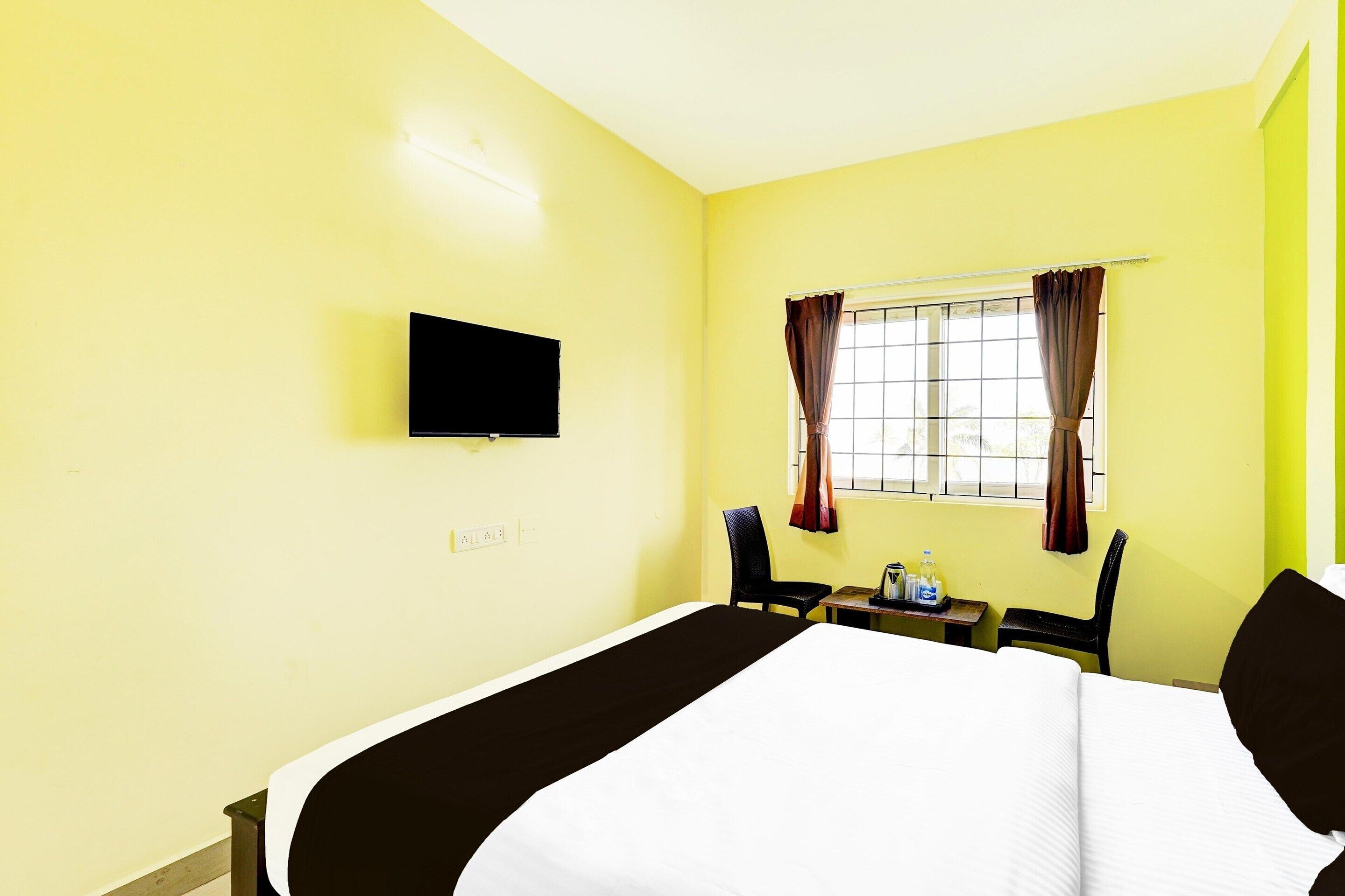 A hotel room with a bed, a television mounted on the wall, a window with curtains, and a small table with a chair.