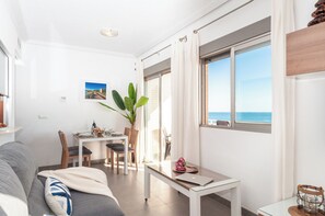 A modern living room with a sofa, a dining table, chairs, a large window with a view of the sea, and a potted plant. - Apartamentos Turisticos Rent Guardamar (Guardamar del Segura)