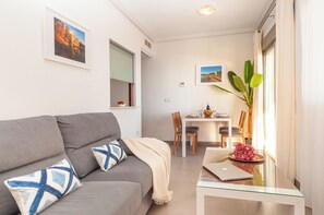 A modern living room with a sofa, a glass table, and a dining area in the background. - Apartamentos Turisticos Rent Guardamar (Guardamar del Segura)