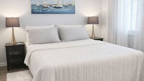 Un dormitorio con una cama grande, dos mesitas de noche, un buró, un cuadro de veleros y una ventana con cortinas.