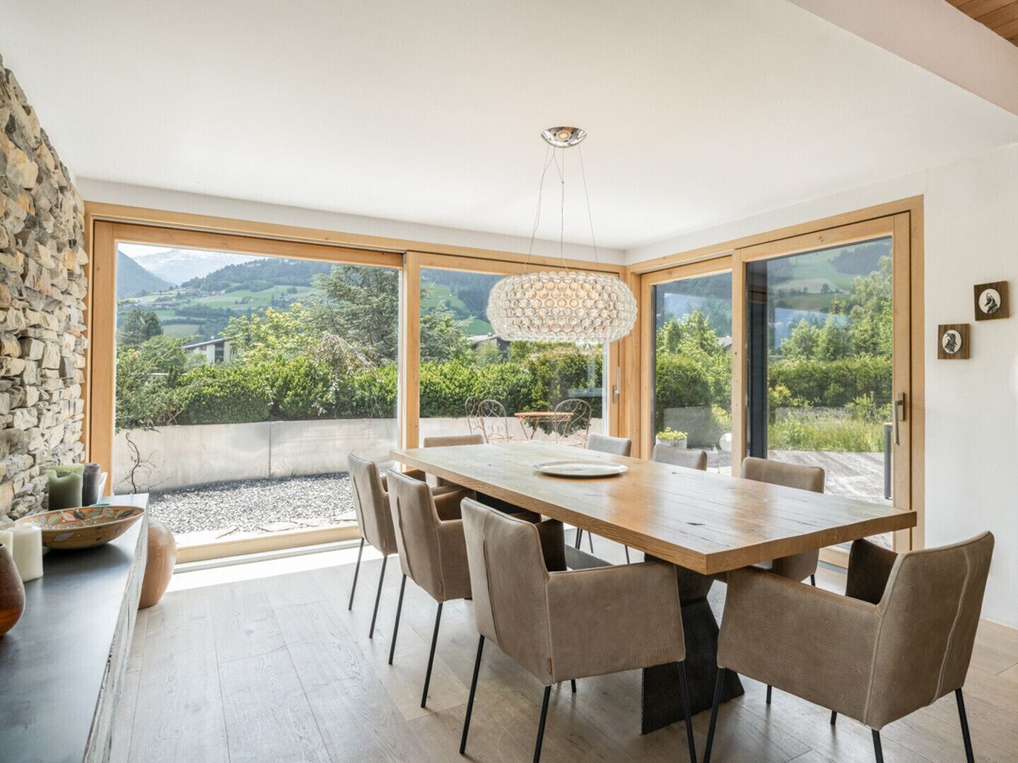 Ein Essbereich mit einem großen Tisch, Stühlen, einer Hängeleuchte und Blick auf Grünflächen und Berge durch große Fenster.