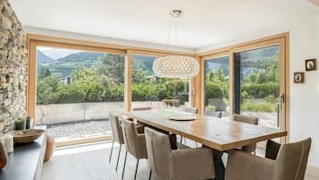 Ein Essbereich mit einem großen Tisch, Stühlen, einer Hängeleuchte und Blick auf Grünflächen und Berge durch große Fenster.