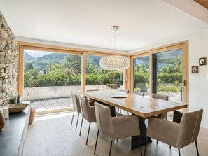 A dining area with a large table, chairs, a hanging light fixture, and a view of greenery and mountains through large windows.