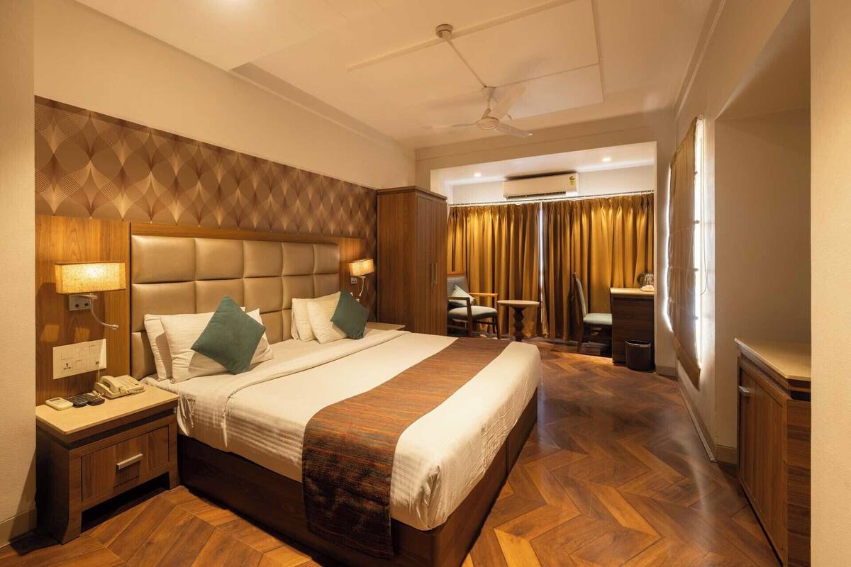 A hotel room with a large bed, a wooden headboard, a desk with a chair, a small table, and a window with curtains.