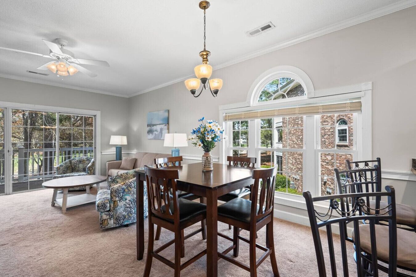 A dining area with a table, chairs, and a chandelier. There is a sofa and a window with a view of a brick building.