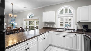 Una cocina con gabinetes blancos, encimera de granito, un lavamanos y un comedor con mesa y sillas.