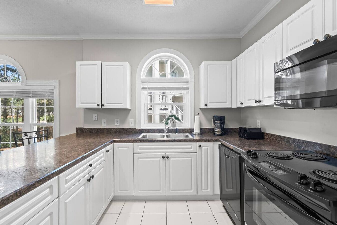 Una cocina con armarios blancos, encimera de granito, estufa de acero inoxidable y ventana con vista a árboles.