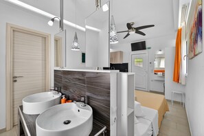 Ein modernes Badezimmer mit zwei Waschbecken, einem Spiegel, Pendelleuchten, einem Deckenventilator und einem Bett mit einer beigen Tagesdecke.