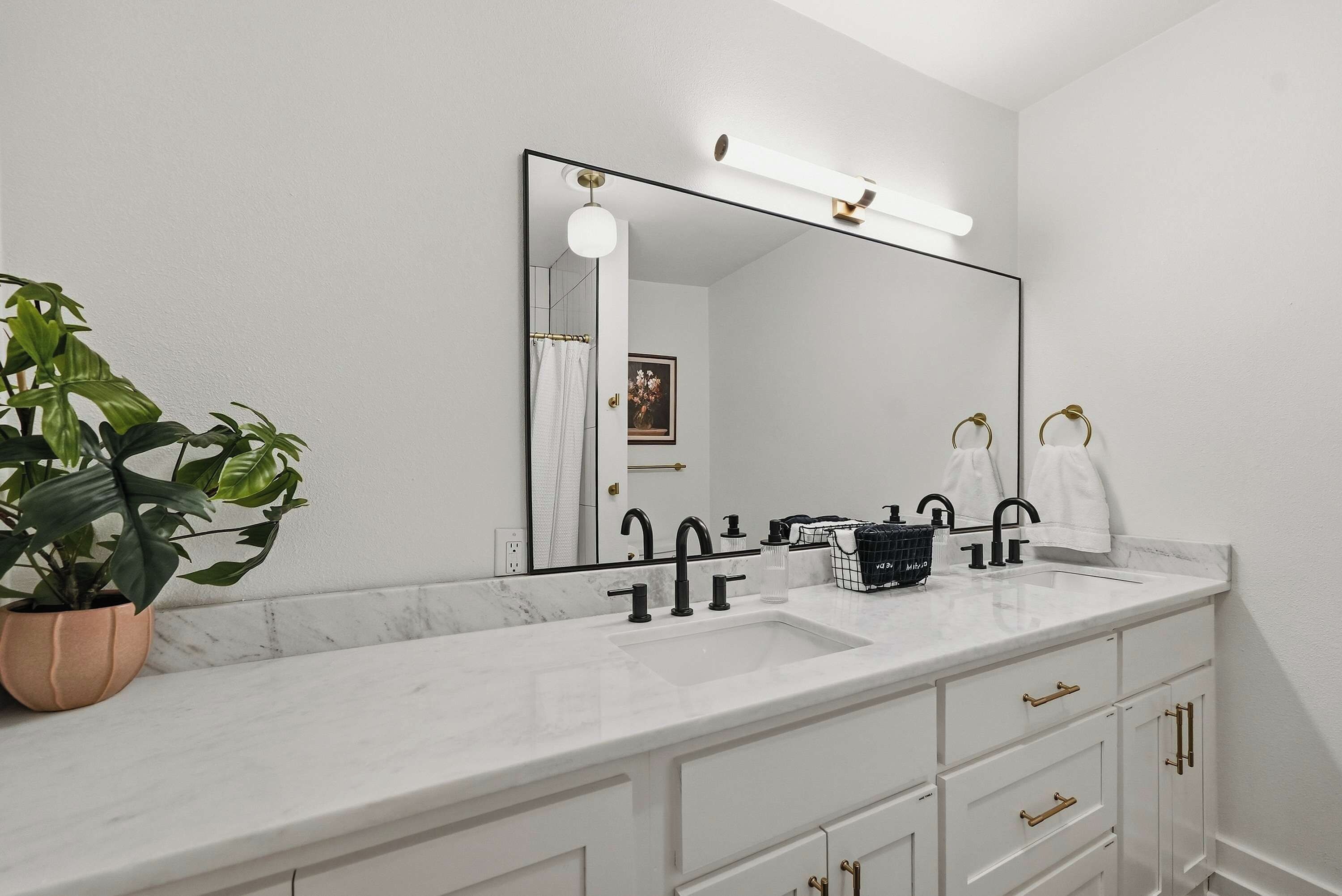 A bathroom with a white marble vanity, black faucets, a large mirror with a light above it, and a potted plant.