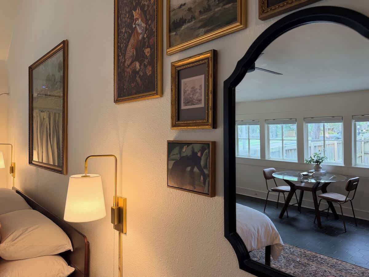 A hotel room with a bed, a lamp, framed pictures on the wall, and a dining area with a table and chairs.