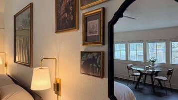 A hotel room with a bed, a lamp, framed pictures on the wall, and a dining area with a table and chairs.