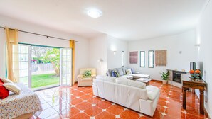 Ein geräumiges Wohnzimmer mit einer großen Couch, einem kleinen Sessel, einem Esstisch und einer Glastür, die in einen Garten führt.