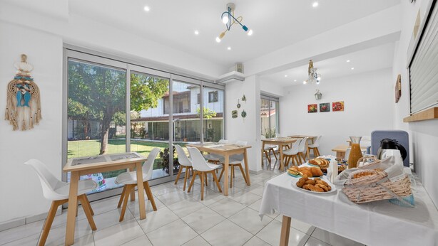 Une salle à manger lumineuse, dotée de grandes fenêtres, de chaises blanches et d’une table dressée avec du pain et des viennoiseries.