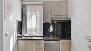 Une cuisine compacte équipée d’un lave-vaisselle, d’une fenêtre agrémentée de rideaux légers et d’un crédence noire.