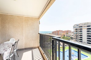 A balcony with a table and chairs, a pool, and a view of buildings and the sea.