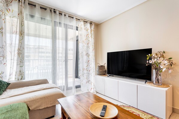 A modern living room with a sofa, a wooden coffee table, a TV on a white cabinet, and a window with floral curtains.
