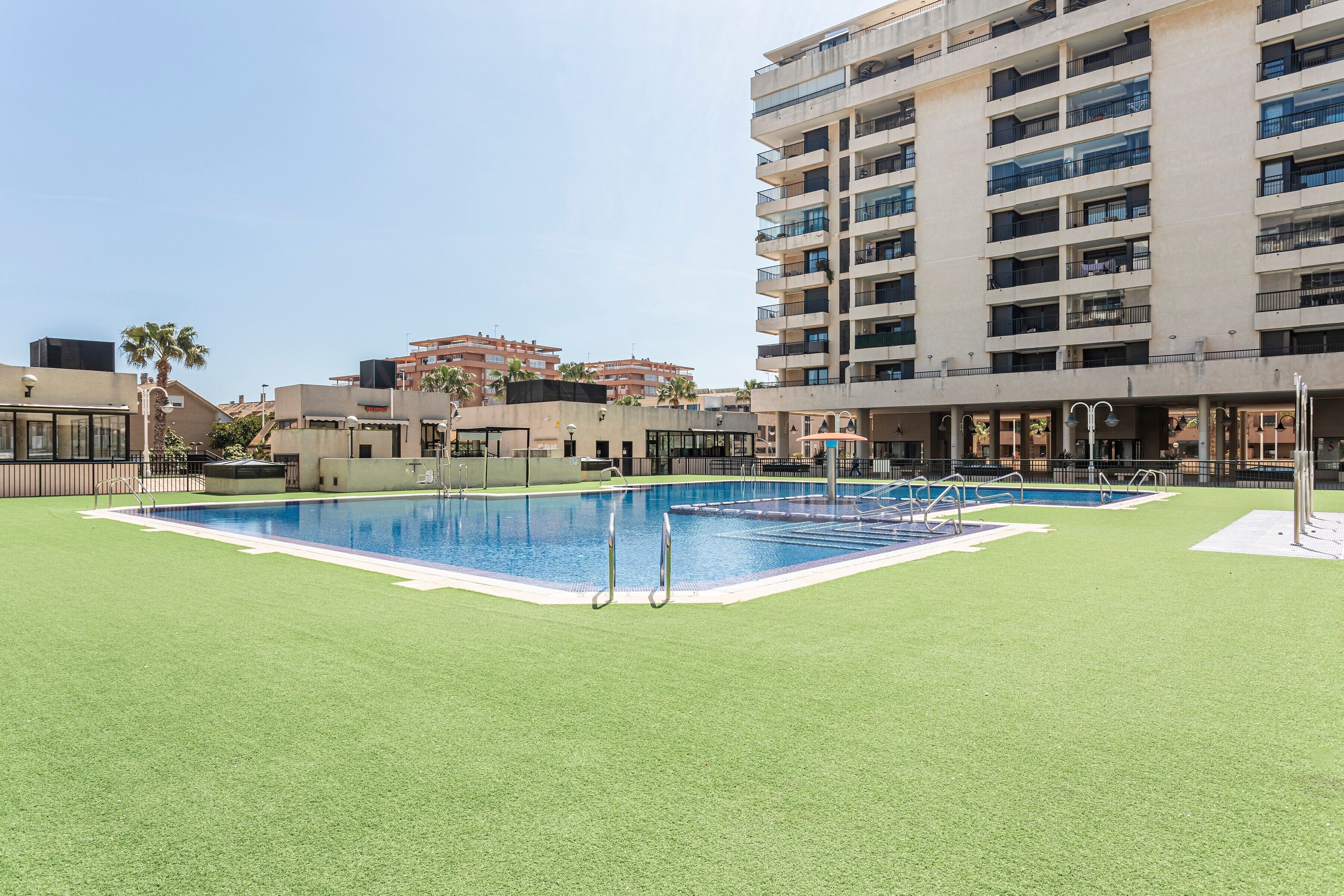 Una piscina con césped verde alrededor, un edificio alto con balcones y un cielo despejado.