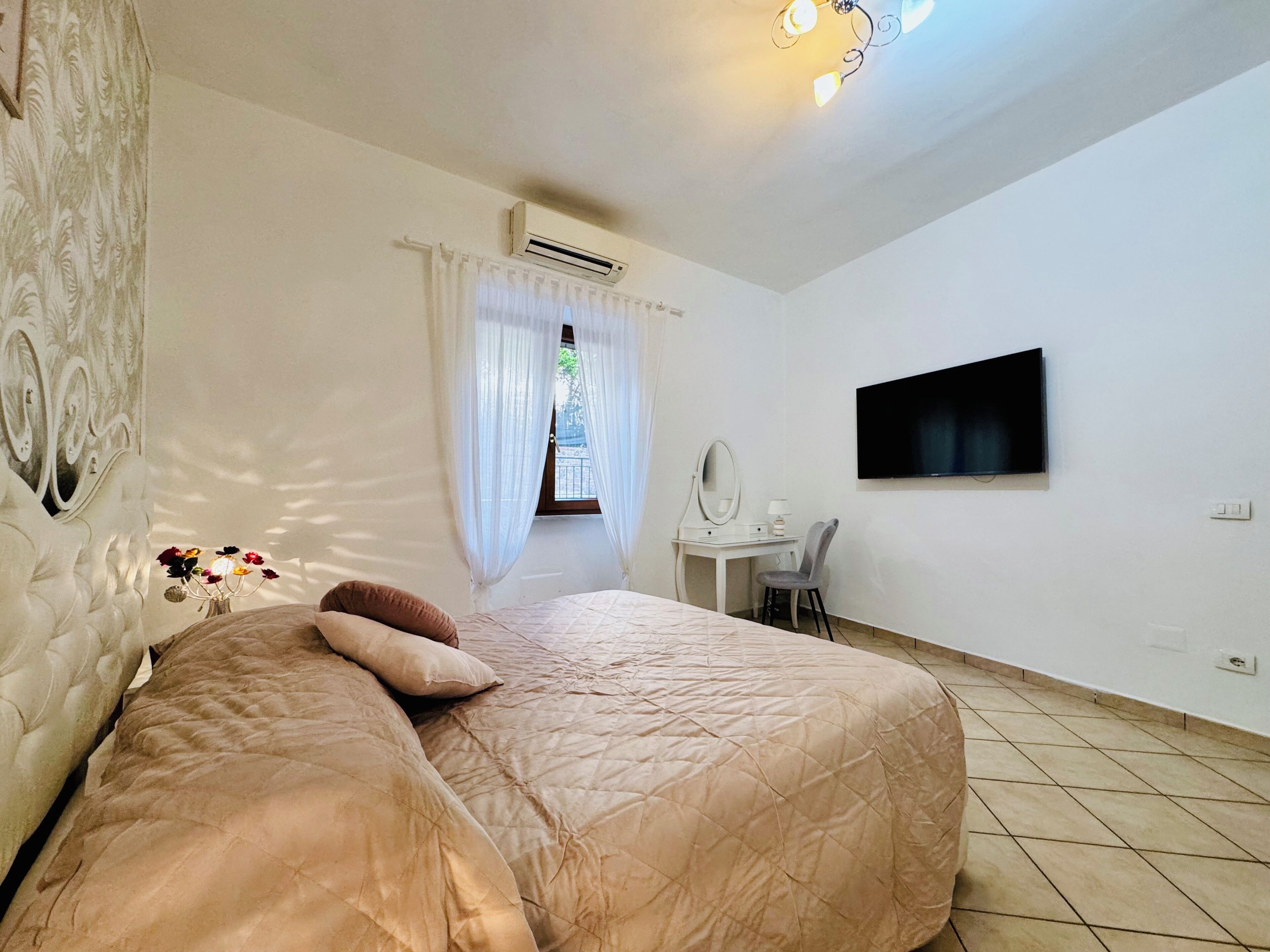 A bedroom with a bed, a desk with a chair, a TV, and an air conditioning unit.