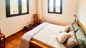 Un dormitorio con cama, dos ventanas, una silla, una mesita de noche con lámpara y una pequeña cesta.