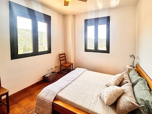 Un dormitorio con cama, dos ventanas, una silla, una mesita de noche con lámpara y una pequeña cesta.