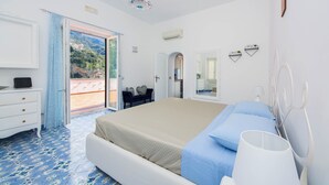 A bedroom with a bed, a dresser, a mirror, and a view of a mountainous landscape through an open door. - YourHome - Lorena Maria House Positano (Positano)