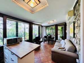 A modern kitchen with a microwave, dining table, and chairs, large windows with a view of greenery, and a stone wall. - YourHome - Maison Iolanda Positano (Positano)
