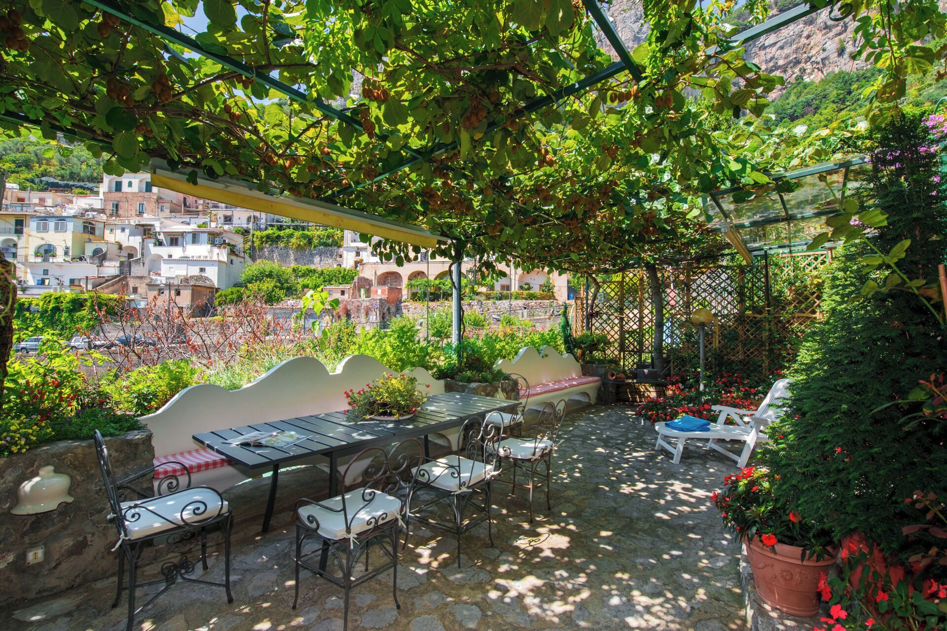 An outdoor patio with stone flooring, metal chairs, a table, and a pergola with climbing plants.
