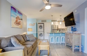 A living room with a sofa, a coffee table, a ceiling fan, a washing machine, a kitchen area, and a TV mounted on the wall. - Walk to Beach, Indoor/Outdoor Pools, Steps to Grocery (Ocean City)