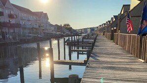 En træbro strækker sig ud i roligt vand, med en klar himmel og solen lavt på horisonten.