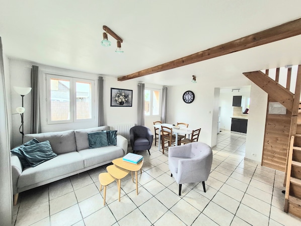 Ein Wohnzimmer mit einer Couch, Sesseln, einem kleinen Tisch, einem Esstisch mit Stühlen, einer Uhr und einer Treppe.