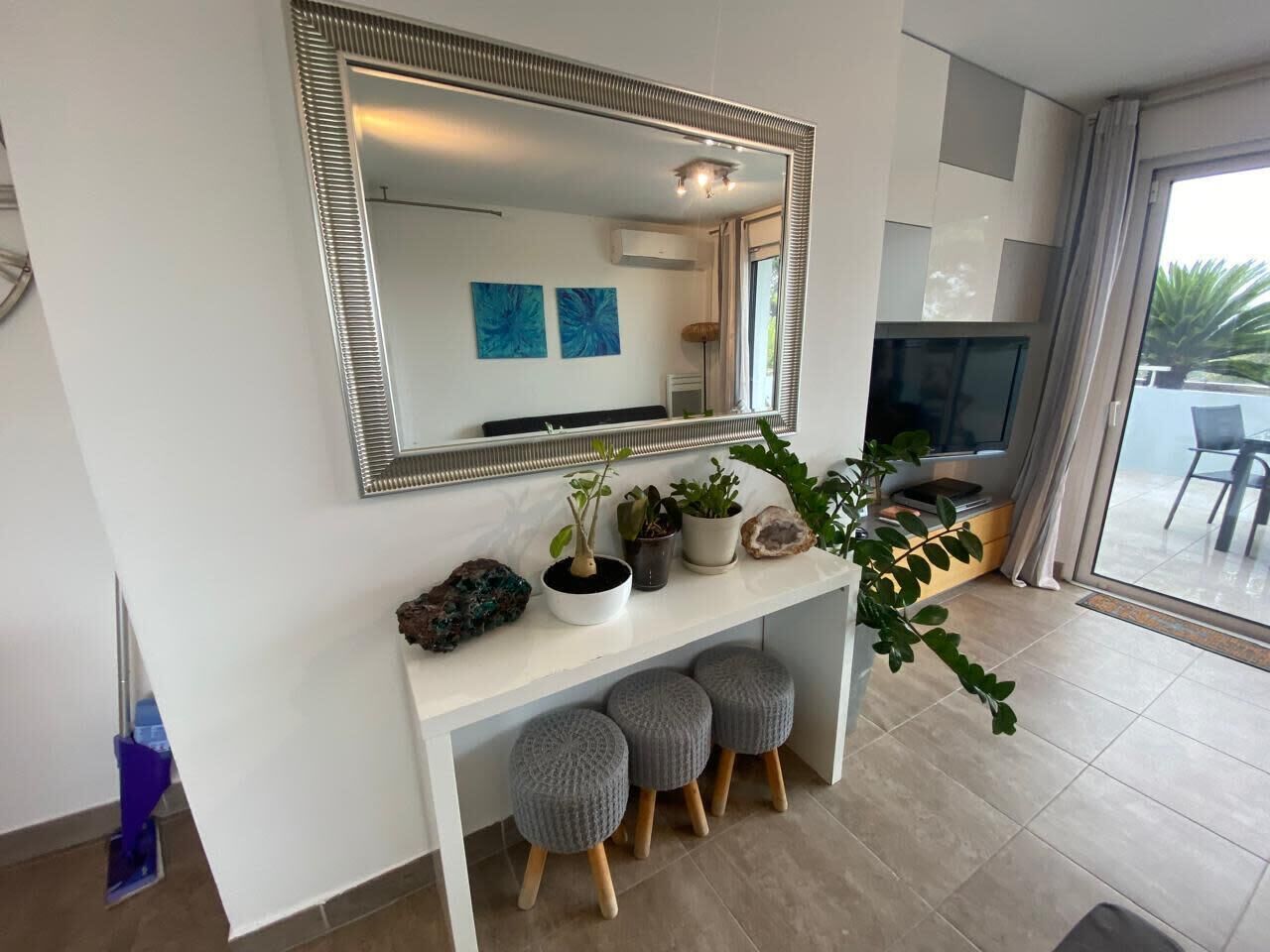 A modern living room with a large mirror, a white console table with potted plants, grey ottomans, and a TV.