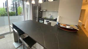 A modern kitchen with a large island, pendant lights, stainless steel appliances, and a view through a glass door.