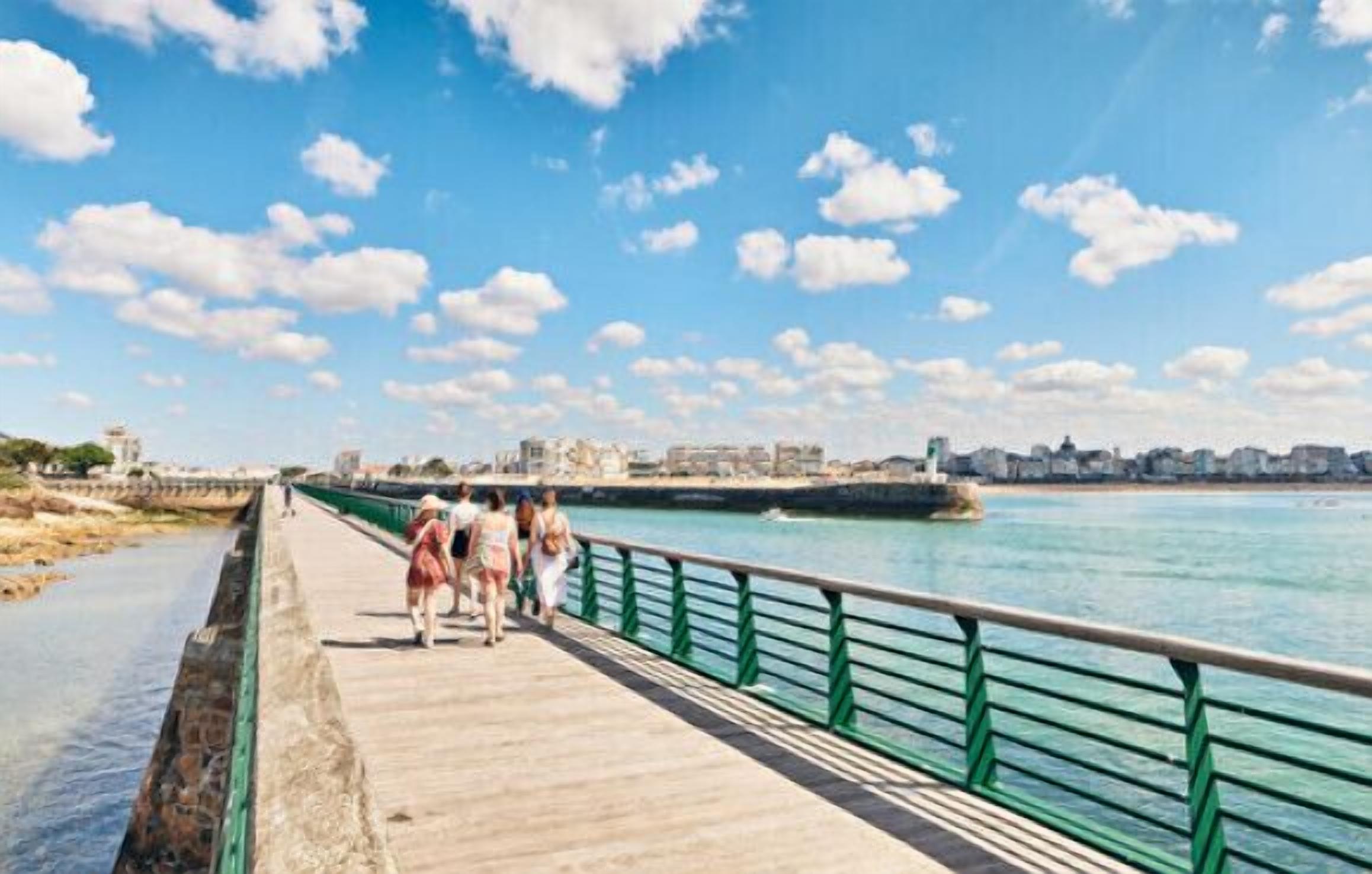 A wooden boardwalk with a green railing alongside a body of water, leading to a cityscape with historic buildings.