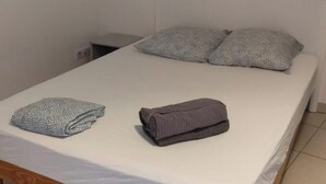 A neatly made bed with a white mattress and two pillows, a folded blanket, and a rolled-up towel on a wooden frame bed in a room with tiled flooring and a patterned wall. - Quiet little house in Bayonne (Bayonne)